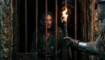 Középkori börtönrácsok mögött sínylődő alak a budavári Labirintus sötétjében, fáklyafénynél.